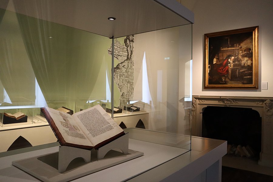 Vitrine mit einem aufgeschlagenen Buch, dahinter ein grüner Vorhang und ein Gemälde über dem Kamin