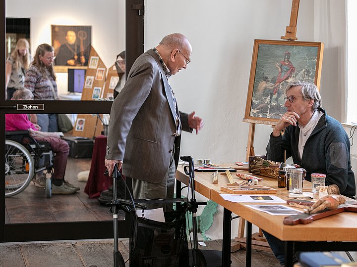 Ein Besucher im Gespräch mit einem Restaurator, daneben ein Gemälde auf der Staffelei