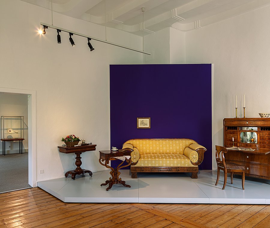 Wohnzimmer-Arrangement mit Sofa und Sekretär vor einem lila Wandfeld