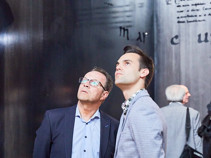 Zwei Besucher blicken an der beschrifteten Stahlwand des Evangeliarraums empor