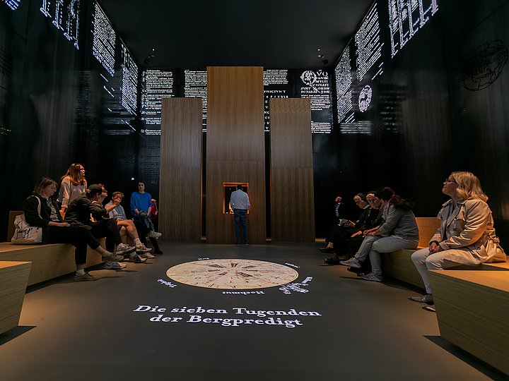 Besucher sitzen im Evangeliarraum, im Boden die sieben Tugenden der Bergpredigt